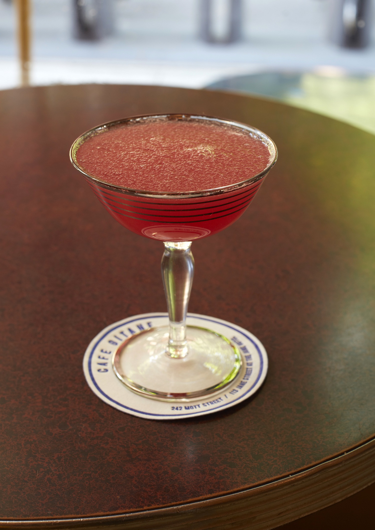 A deep red cocktail in a coupe glass on a dark table