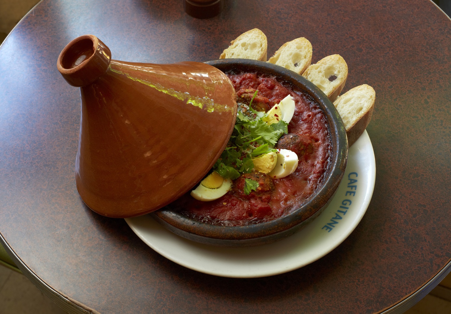 A terracotta tagine beside a dish garnished with egg and sliced baguette