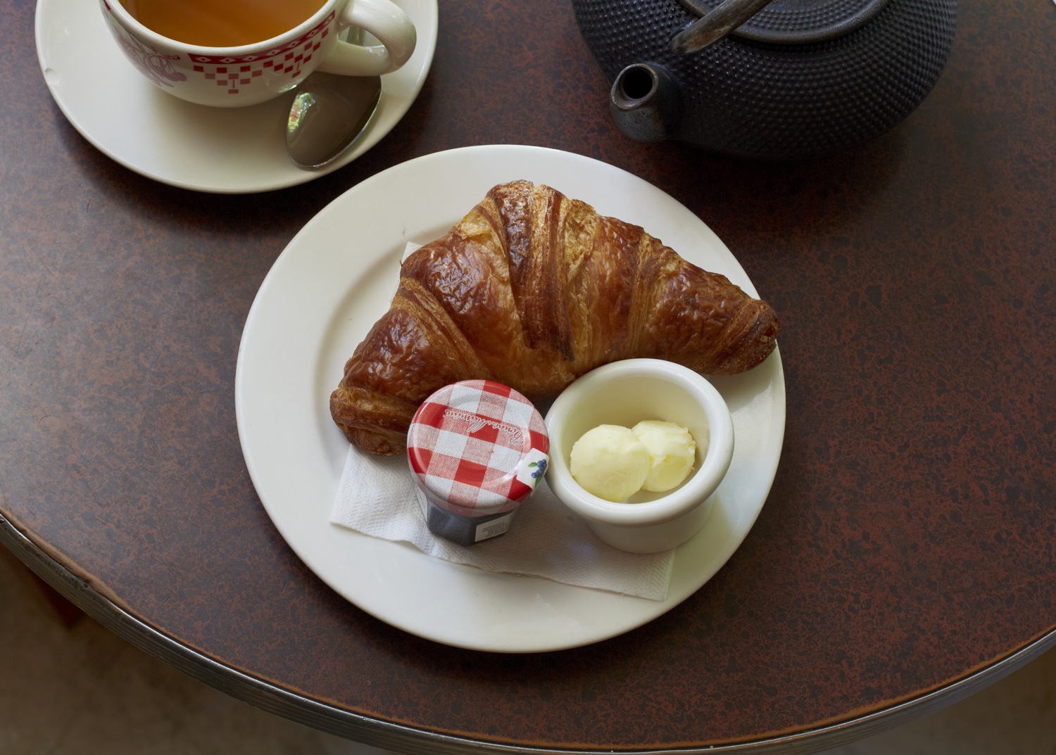 A croissant with butter and jam beside a cup of coffee and a teapot